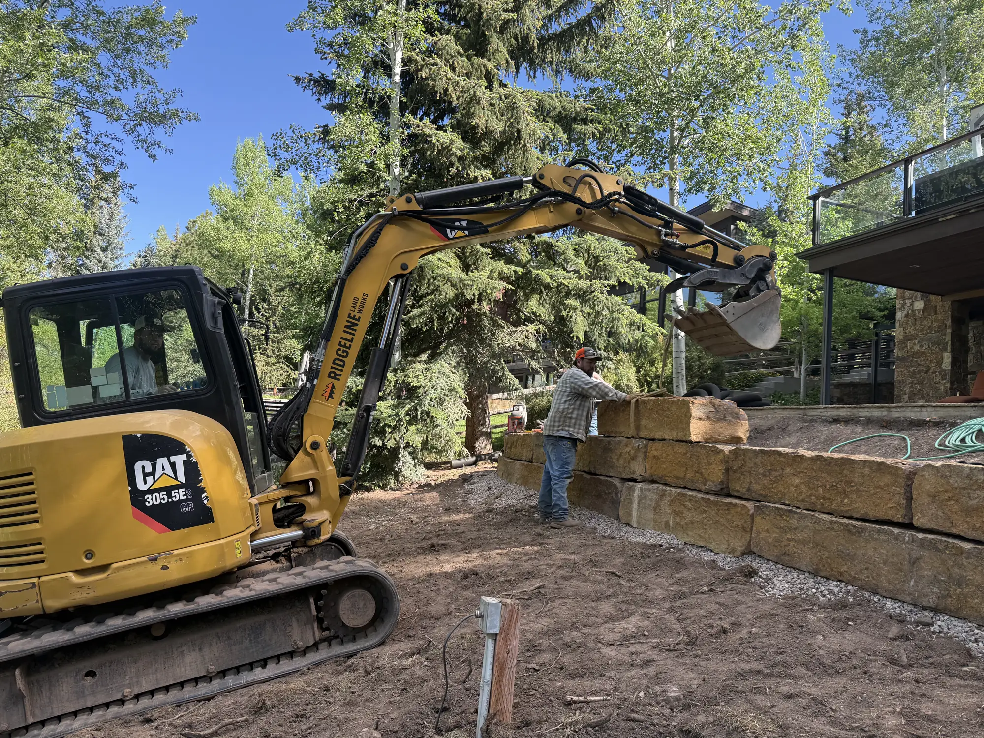 Excavator setting a sandstone block retaining wall with a crew member guiding the block