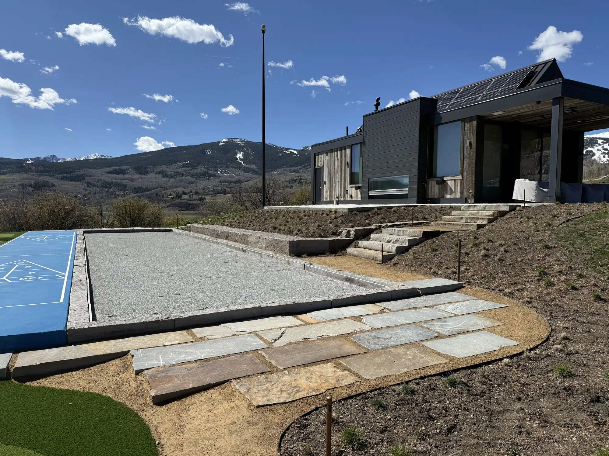 Bocce court, flagstone patio and stone retaining walls built into a hillside below a modern home with mountains beyond
