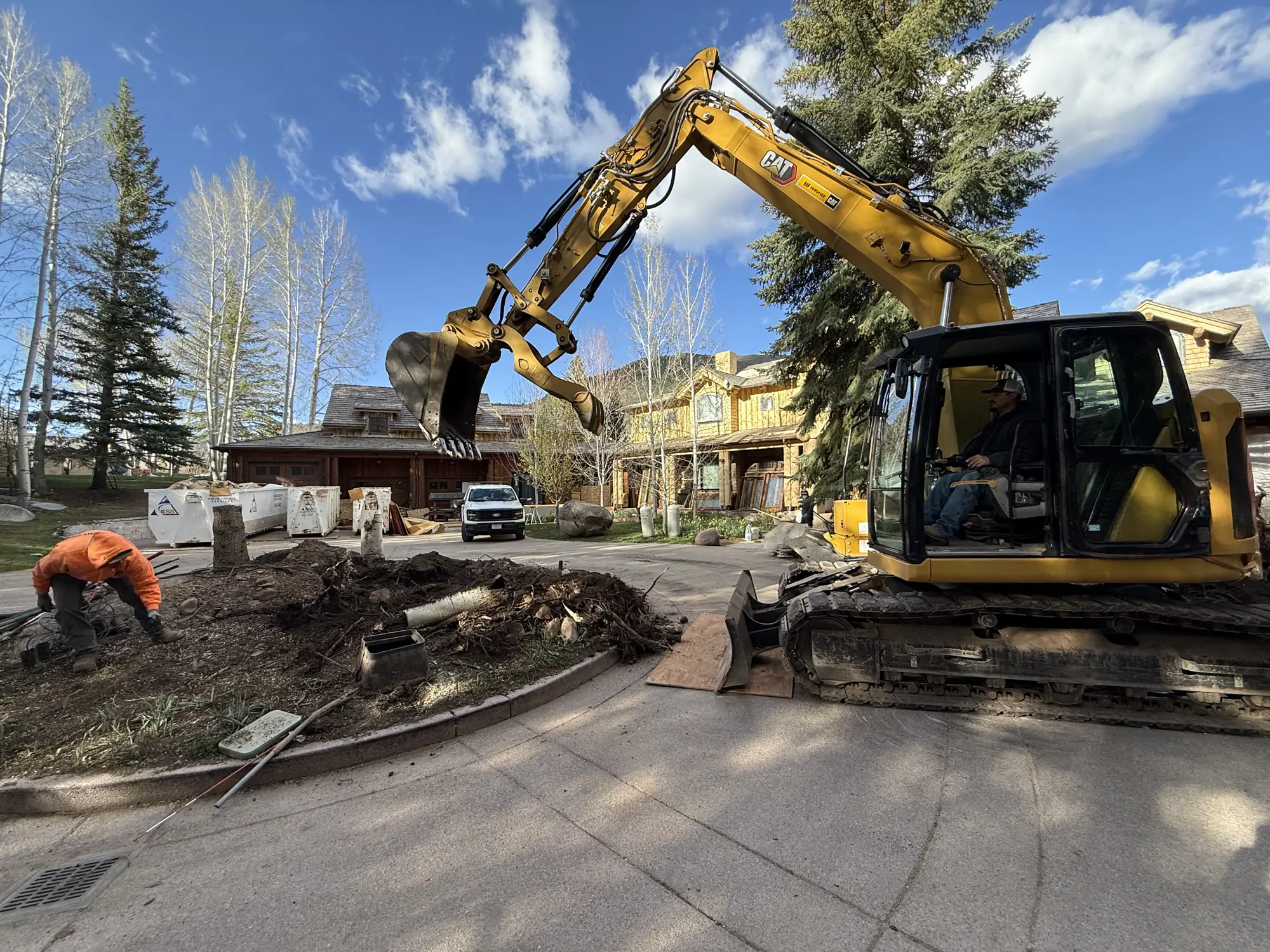 Excavator removing a driveway island while a crew member works alongside